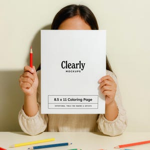 May include: A white 8.5 x 11 coloring page with the text "Clearly Mockups" and "Intentional Tools for Makers & Artists". A red pencil is held in one hand. Several colored pencils are on the table.