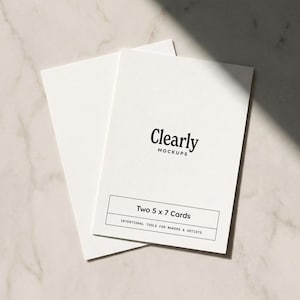 May include: Two 5 x 7 inch white cards with the text "Clearly Mockups" and "Two 5 x 7 Cards" in black. The cards are arranged on a light-colored surface, with a shadow across the top.