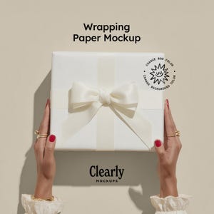May include: A white gift box wrapped in white paper and tied with a satin bow and ribbon. The text "Wrapping Paper Mockup" is at the top, and "Clearly Mockups" is at the bottom. The hands holding the box have red nail polish and gold rings.