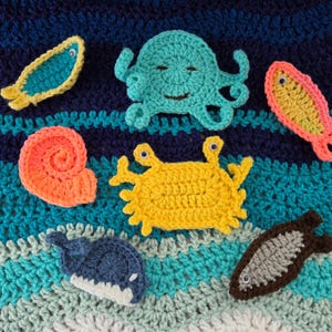 May include: A collection of crocheted sea creatures including a teal octopus, a yellow crab, a coral seashell, a blue whale, and two small fish in yellow and brown, all featuring small plastic eyes and arranged on a blue and teal crocheted background.
