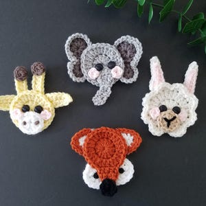 May include: Four crocheted animal face appliques: a yellow giraffe, a gray elephant, a white bunny, and an orange fox. Each applique features button eyes and pink accents. The giraffe has brown spots, and the fox has a white face.
