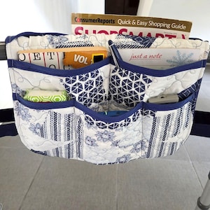 May include: A quilted walker bag featuring a white and blue geometric and floral pattern with dark blue trim. The bag has multiple exterior pockets containing a Consumer Reports ShopSmart magazine, a book, a notepad labeled just a note, and a pen.
