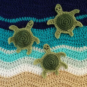 May include: Three crocheted sea turtles in shades of olive and forest green are positioned on a crocheted background featuring wavy horizontal stripes of dark blue, teal, light blue, white, and tan, mimicking an ocean and sandy beach scene.