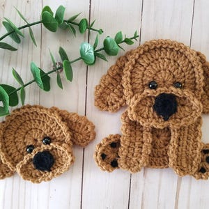 May include: Two brown crocheted dog figures made of yarn. The smaller figure is a dog head with black eyes and a black nose. The larger figure is a full-body dog with floppy ears, black eyes, a black nose, and black paw pads on its front feet.