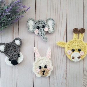 May include: Crocheted animal faces including a gray elephant, a yellow giraffe, a gray koala, and a white bunny with pink accents. The handmade items are arranged on a light wood surface with a sprig of purple flowers.