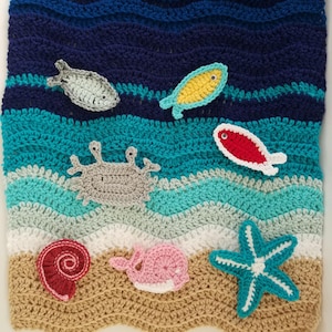 May include: A crocheted blanket featuring a wavy ocean pattern in shades of dark blue, teal, light blue, white, and tan. Appliqued sea creatures include a gray crab, a yellow fish, a red fish, a gray fish, a red shell, a pink whale, and a teal starfish.