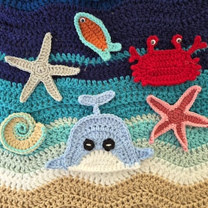 May include: A crochet blanket featuring a wavy ocean pattern in shades of dark blue, teal, and light blue, transitioning to a tan sandy base. Appliqués include a beige starfish, an orange fish, a red crab, a spiral shell, and a light blue whale.