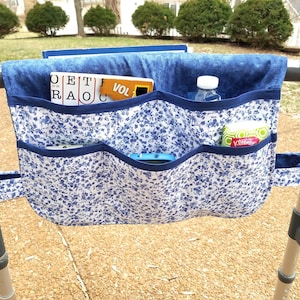 May include: A blue and white floral print walker bag with multiple pockets, attached to a gray walker. The bag holds a book, a water bottle, tissues, and other items. The bag is made of fabric with a blue floral pattern.