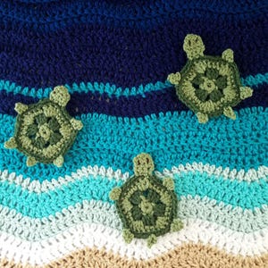May include: A crocheted blanket featuring a wavy pattern in shades of dark blue, teal, light blue, white, and tan. Three small, green, crocheted sea turtle appliques are attached to the surface, positioned across the blue and teal sections.
