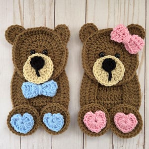 May include: Two brown crocheted teddy bear appliques. The left bear features a light blue bow tie and matching light blue heart-shaped foot pads. The right bear features a pink bow on its head and matching pink heart-shaped foot pads.