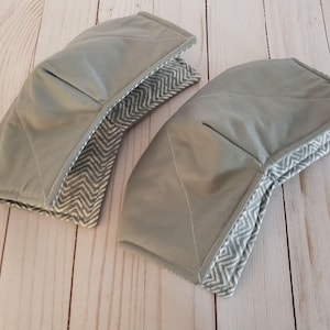 K&ouml;nnte beinhalten: Zwei graue Topflappen aus Stoff mit einem wei&szlig;en und grauen Zickzackmuster. Die Topflappen sind aus weichem, gestepptem Stoff.