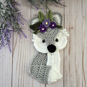 May include: A crochet fox applique featuring a gray and white color scheme with black bead eyes and a black nose. The fox is adorned with a crown of crocheted green leaves and small purple flowers with yellow centers, crafted with detailed yarn texture.
