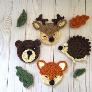 May include: A collection of crochet appliques featuring a brown deer, a dark brown bear, a brown hedgehog, and an orange fox, accompanied by two green leaves and one orange oak leaf, all crafted with detailed yarn stitching and distinct facial features.