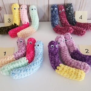 May include: A collection of handmade, plush worm toys in various colors. Each worm has a fuzzy texture, googly eyes, and is segmented with different color combinations, including pink, blue, yellow, and purple. The toys are arranged on a white surface.