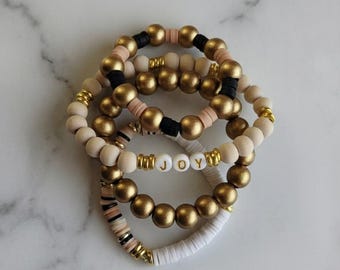 May include: A collection of beaded bracelets in various styles. The bracelets feature gold, white, black, and tan beads. One bracelet spells out the word "JOY" with white letter beads. The bracelets are stacked together.