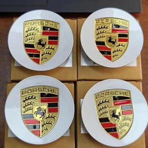 May include: Four white and gold Porsche wheel center caps. Each cap features the Porsche crest in the center, with a black horse, gold accents, and red and black stripes. The caps are round and sit on cardboard boxes.