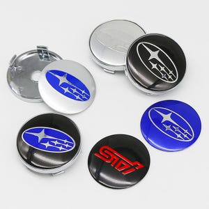 May include: A collection of circular wheel center caps featuring the Subaru logo in blue, black, and silver, along with one black cap displaying the red STI logo. These automotive accessories have a glossy finish and metallic backing components.