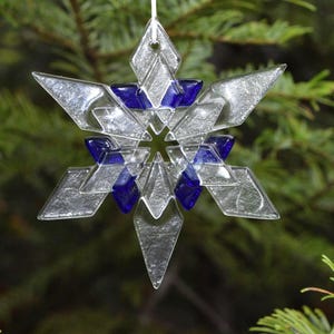 May include: A clear glass snowflake ornament with blue accents hangs from a white string against a backdrop of green pine needles. The ornament has a geometric design with six points and is suitable for holiday decor.