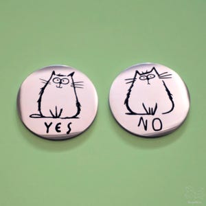 May include: Two round, silver-colored buttons with black cartoon cat illustrations. One button says "YES" and the other says "NO". The cats have surprised expressions and are drawn with thick black lines.