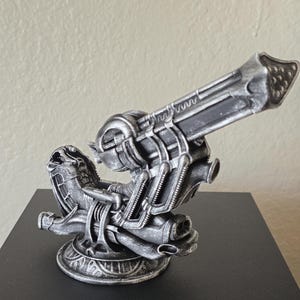 May include: A detailed, silver-colored model of a futuristic cannon. The design features intricate mechanical details, including ribbed barrels and a complex base. The cannon is positioned at an angle, suggesting a dynamic pose.