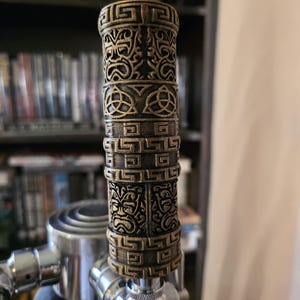 May include: A decorative beer tap handle with an intricate, aged bronze design. The handle features detailed patterns and shapes, including Celtic knots and stylized faces. The tap is attached to a stainless steel faucet.