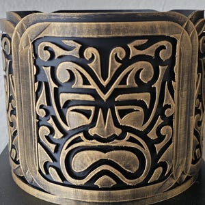 May include: A cylindrical decorative container with a black base and a detailed, ornate design. The exterior features a repeating pattern of a stylized face and swirling motifs in a bronze color, creating a striking contrast against the black background.