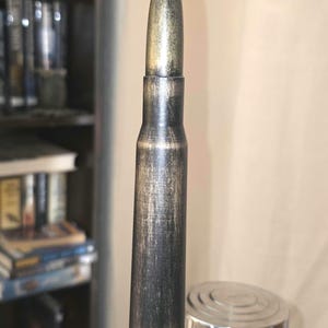 Puede incluir: Objeto metálico con forma de bala, con punta dorada y cuerpo oscuro y texturizado. Montado sobre una base plateada, sugiere un uso decorativo o funcional. El diseño general evoca un estilo vintage o industrial.