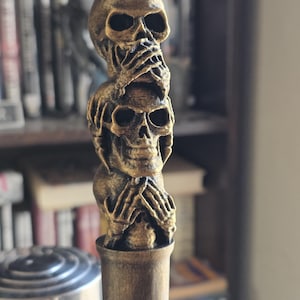 May include: A gold-colored bottle stopper featuring three stacked skulls, each with hands covering their eyes, mouth, and ears. The stopper has a cylindrical base and is designed with intricate details, creating a unique decorative piece.