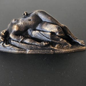 May include: A bronze-colored sculpture of a reclining figure. The figure is lying on a textured base, with details in the hair and body. The sculpture has a dark bronze color with gold highlights.