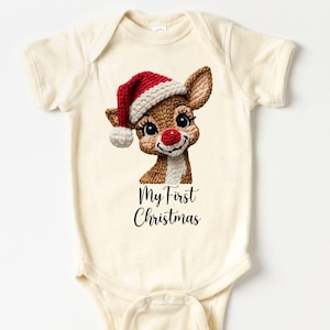 May include: Cream-colored baby onesie featuring a cartoon reindeer wearing a red and white Santa hat. The reindeer has a red nose and is smiling. The text "My First Christmas" is printed below the reindeer.