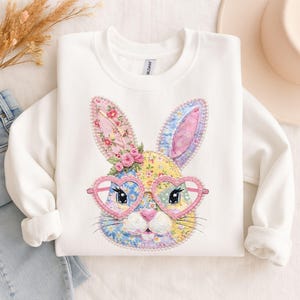 Pode incluir: Camisola branca com um design de coelho de patchwork colorido com óculos em forma de coração. As orelhas do coelho são adornadas com padrões florais e rosas cor-de-rosa. O design é bordado, criando uma aparência texturizada. Uma peça de roupa casual e festiva.