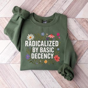 May include: An olive green sweatshirt with the text "RADICALIZED BY BASIC DECENCY" in white, surrounded by a floral design with daisies, pink, blue, and yellow flowers. The sweatshirt has long sleeves.