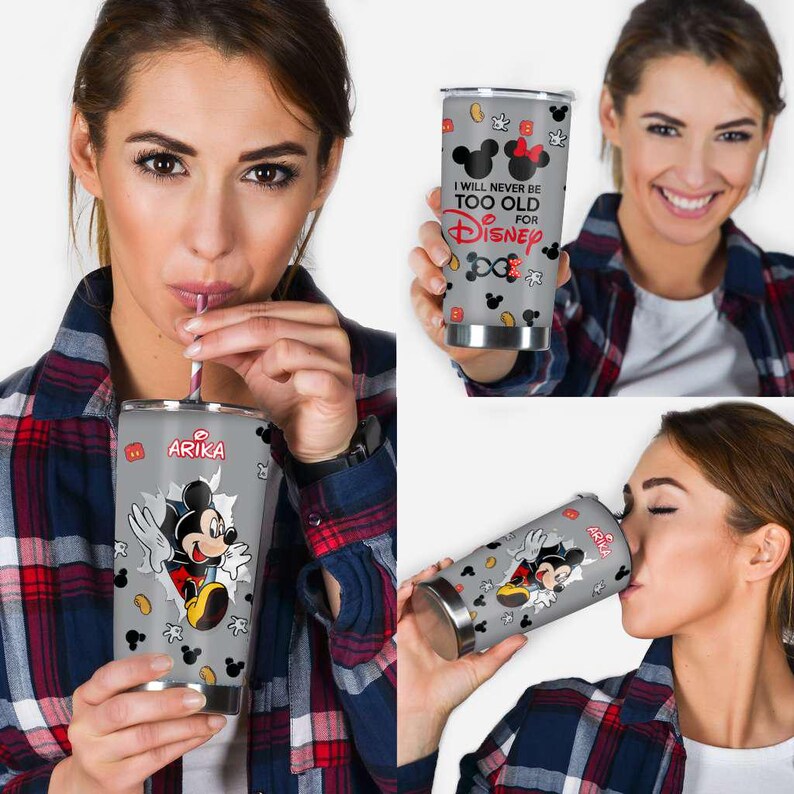 May include: A grey stainless steel tumbler with a Mickey Mouse design. The tumbler has the name "Arika" and the phrase "I will never be too old for Disney" printed on it, along with Mickey Mouse icons. The tumbler has a silver rim.
