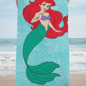 Op de afbeelding: Een strandlaken met een cartoonillustratie van Ariel, de Kleine Zeemeermin, met rood haar, een paarse schelpenbeha en een groene staart. De handdoek heeft een lichtblauwe achtergrond met witte bladerpatronen.