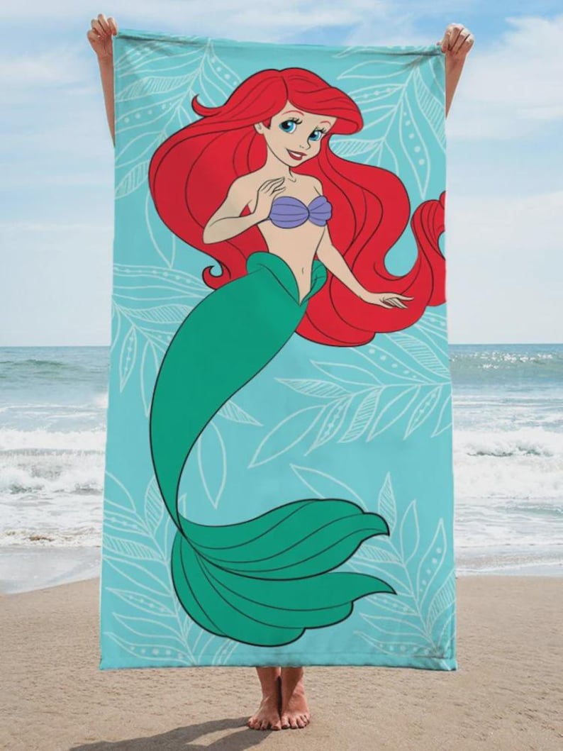 Op de afbeelding: Een strandlaken met een cartoonillustratie van Ariel, de Kleine Zeemeermin, met rood haar, een paarse schelpenbeha en een groene staart. De handdoek heeft een lichtblauwe achtergrond met witte bladerpatronen.