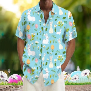 May include: A light blue, short-sleeved button-down shirt featuring a repeating pattern of white rabbits, Easter eggs, and floral elements. The shirt has a classic collar and is made of a lightweight fabric, perfect for spring or summer.