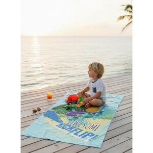 May include: A child sits on a blue beach towel with the words "AWESOME KICKFLIP!" printed on it. The towel has a skateboard graphic. The child is playing with toy trucks and dinosaurs. A glass of orange juice and sunglasses are nearby.