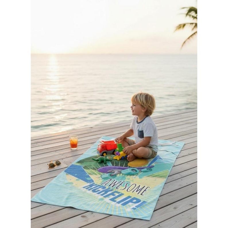 May include: A child sits on a blue beach towel with the words "AWESOME KICKFLIP!" printed on it. The towel has a skateboard graphic. The child is playing with toy trucks and dinosaurs. A glass of orange juice and sunglasses are nearby.