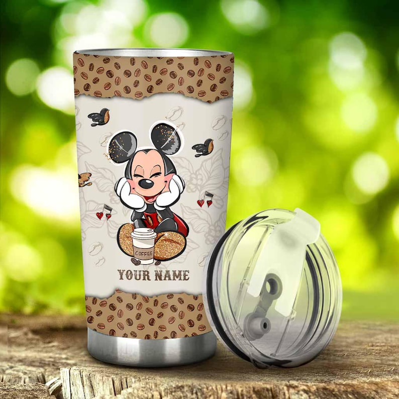May include: A stainless steel tumbler featuring a design of Mickey Mouse with a coffee cup. The tumbler has a brown coffee bean pattern at the top and bottom. The text "COFFEE" and "YOUR NAME" are visible on the tumbler.