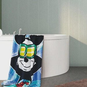 May include: A colorful beach towel featuring Mickey Mouse wearing sunglasses. The towel has a black background with a large image of Mickey's face and a blue and green tropical design. The towel is draped over the edge of a white bathtub.