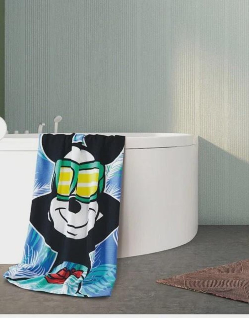 May include: A colorful beach towel featuring Mickey Mouse wearing sunglasses. The towel has a black background with a large image of Mickey's face and a blue and green tropical design. The towel is draped over the edge of a white bathtub.