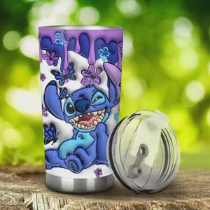 May include: A stainless steel tumbler featuring a cartoon character in shades of blue, with a wide grin and surrounded by butterflies and flowers. The design includes dripping purple paint. The clear lid is placed next to the tumbler.
