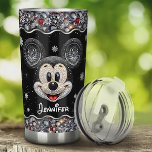 May include: A black tumbler featuring a sparkling Mickey Mouse design with the name "Jennifer" in white script. The tumbler has a silver base and is adorned with decorative gems and snowflakes. A clear lid is next to the tumbler.