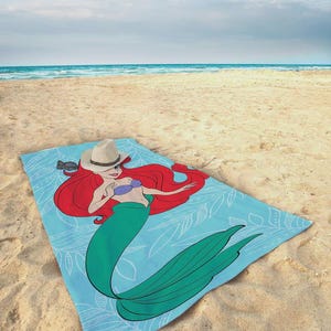 Op de afbeelding: Lichtblauw strandlaken met een cartoonillustratie van een zeemeermin. De zeemeermin heeft rood haar, een groene staart en een paarse top. Een wit bladerpatroon versiert de handdoek en een cowboyhoed ligt erop.