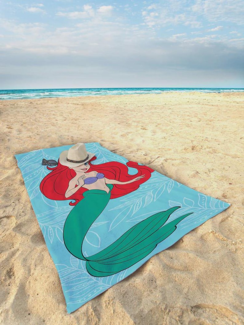 Op de afbeelding: Lichtblauw strandlaken met een cartoonillustratie van een zeemeermin. De zeemeermin heeft rood haar, een groene staart en een paarse top. Een wit bladerpatroon versiert de handdoek en een cowboyhoed ligt erop.