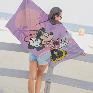 May include: A lavender beach towel featuring Minnie Mouse in sunglasses, a pink polka-dot dress, and yellow shoes. The towel has a sunset and palm tree design. The towel is being held up by a person on a wooden deck.