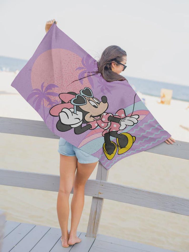 May include: A lavender beach towel featuring Minnie Mouse in sunglasses, a pink polka-dot dress, and yellow shoes. The towel has a sunset and palm tree design. The towel is being held up by a person on a wooden deck.