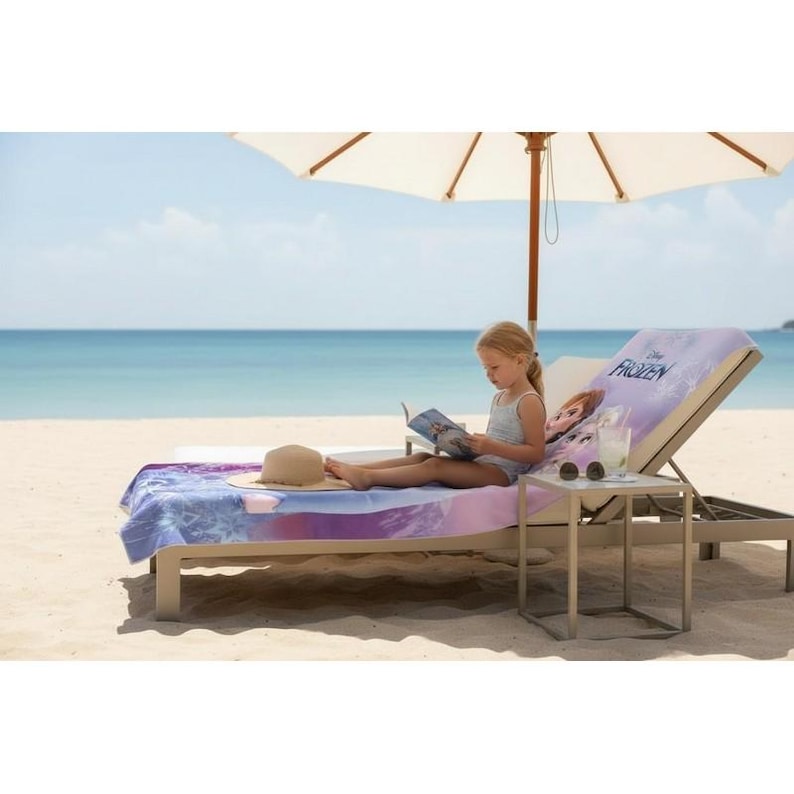 K&ouml;nnte beinhalten: Ein junges M&auml;dchen liest auf einer Sonnenliege am Strand unter einem wei&szlig;en Sonnenschirm. Die Liege hat ein Frozen-Handtuch mit Elsa und dem Frozen-Logo. Ein Strohhut und ein Getr&auml;nk stehen auf einem kleinen Tisch.