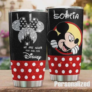 Op de afbeelding: Twee zwarte bekers met zilveren glitterontwerpen. E&eacute;n heeft een Minnie Mouse strik en kasteel, de tekst "WE ARE NEVER TOO OLD FOR Disney". De andere heeft Mickey Mouse en de naam "Sophia". Beide hebben rode stippenbanden.
