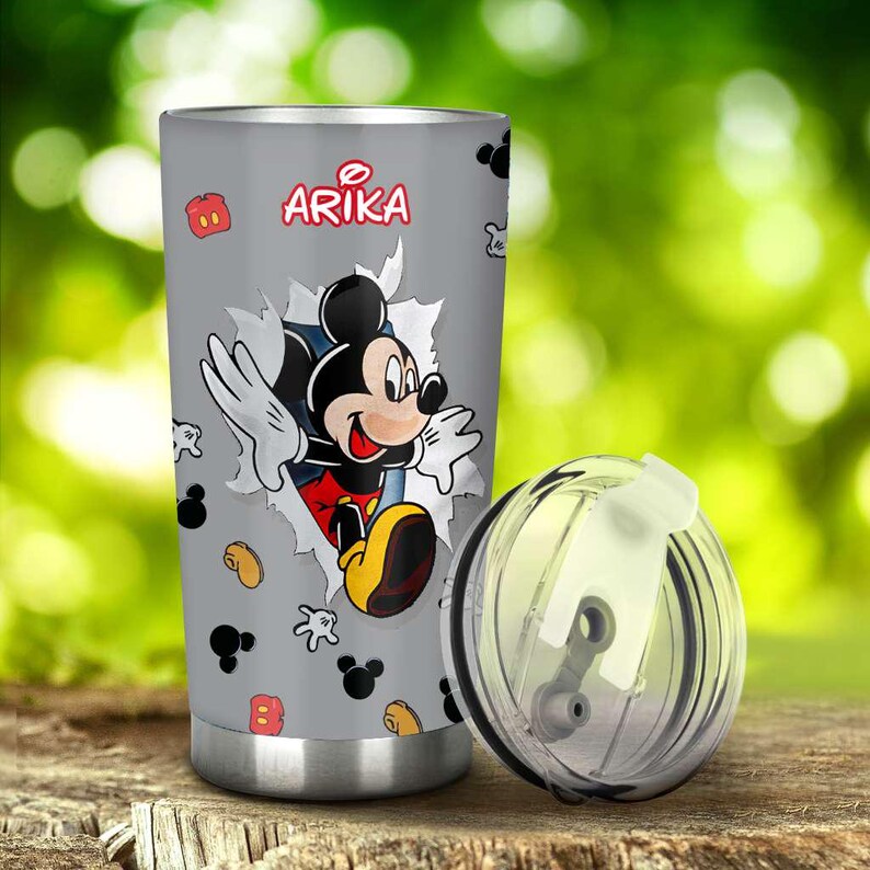 May include: A grey tumbler with a Mickey Mouse graphic breaking through a torn paper design. The name "Arika" is printed in red above the character. The tumbler has a silver base and a clear lid.